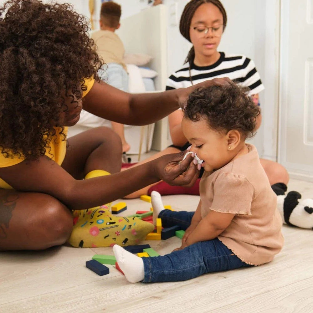 Fürsorgliche Mutter wischt ihrem Baby im Kinderzimmer die Nase; überall liegt Spielzeug verstreut.